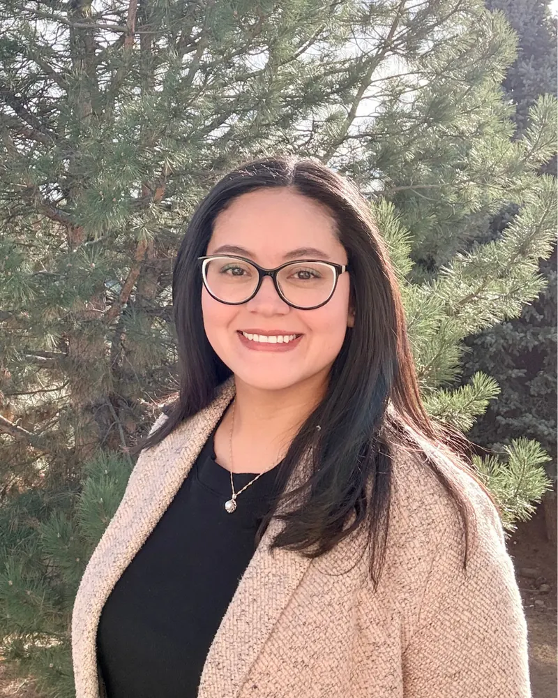 Angela Martinez Lopez, smiling, standing outdoors in front of evergreen trees